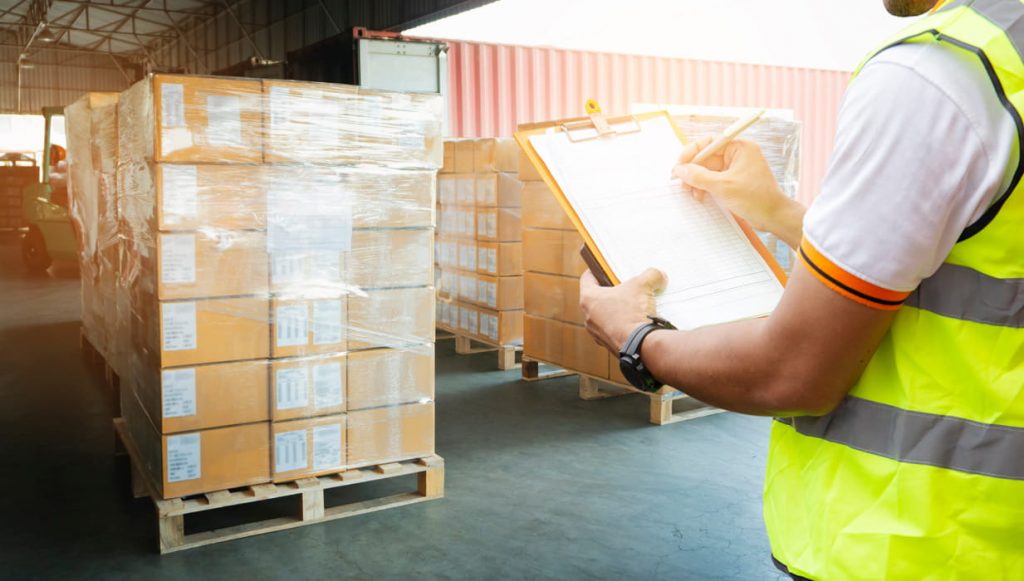 Lagerarbeiter in Warnweste schreibt auf Klemmbrett vor folienverpackten Paletten mit Kartons.