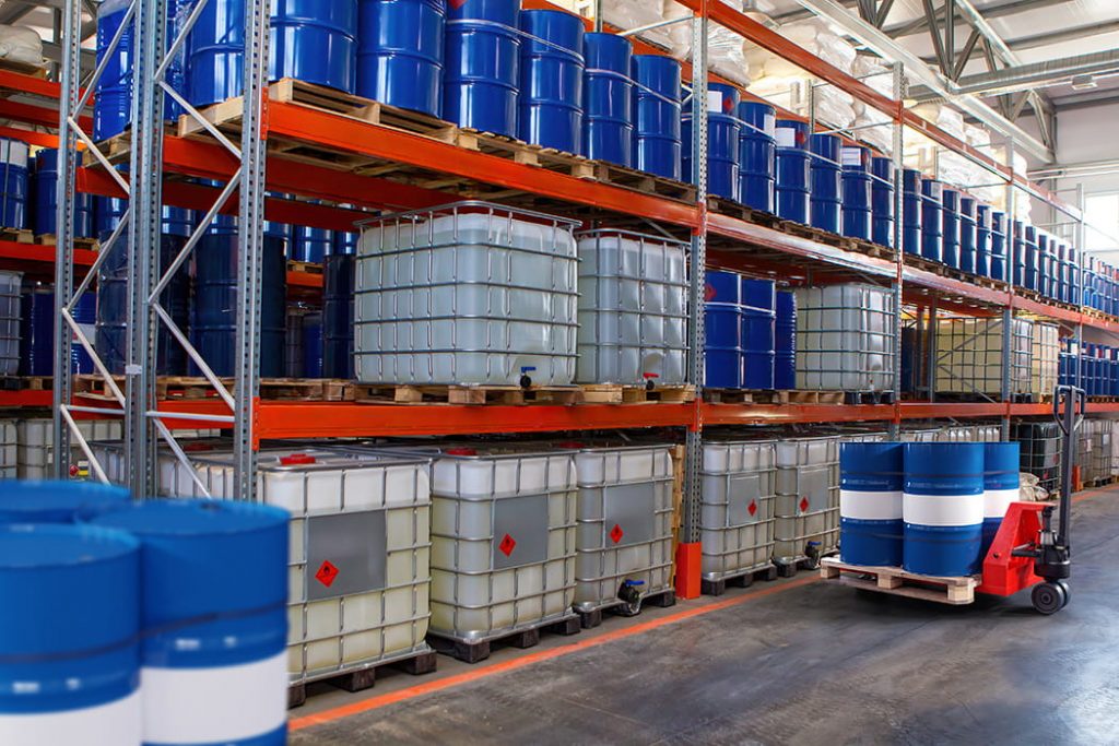 Gefahrstofflager mit beschrifteten IBC-Containern, blauen Fässern und Palettenregalen in der Lagerhalle.