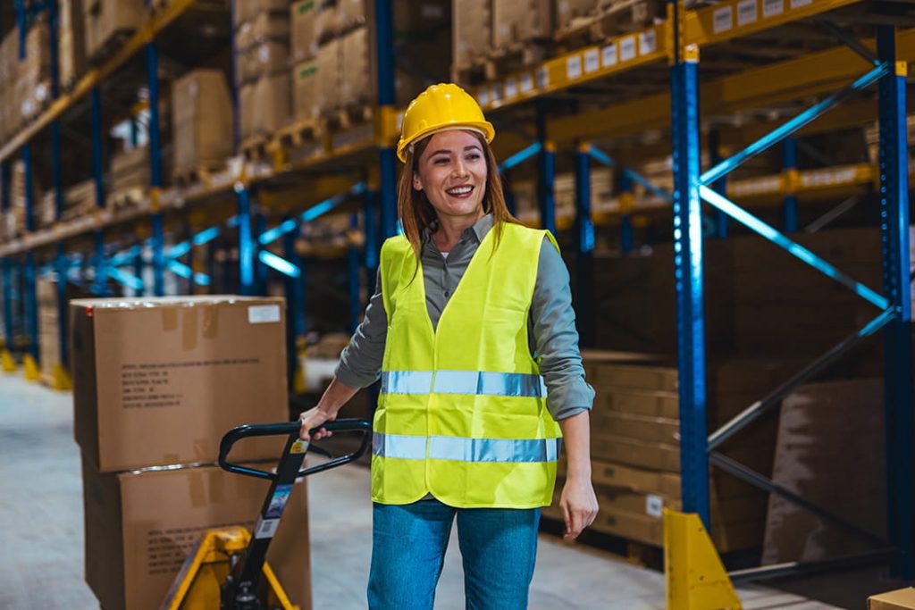Lächelnde Lagermitarbeiterin in Warnschutzweste zieht Hubwagen in Lagerhalle.