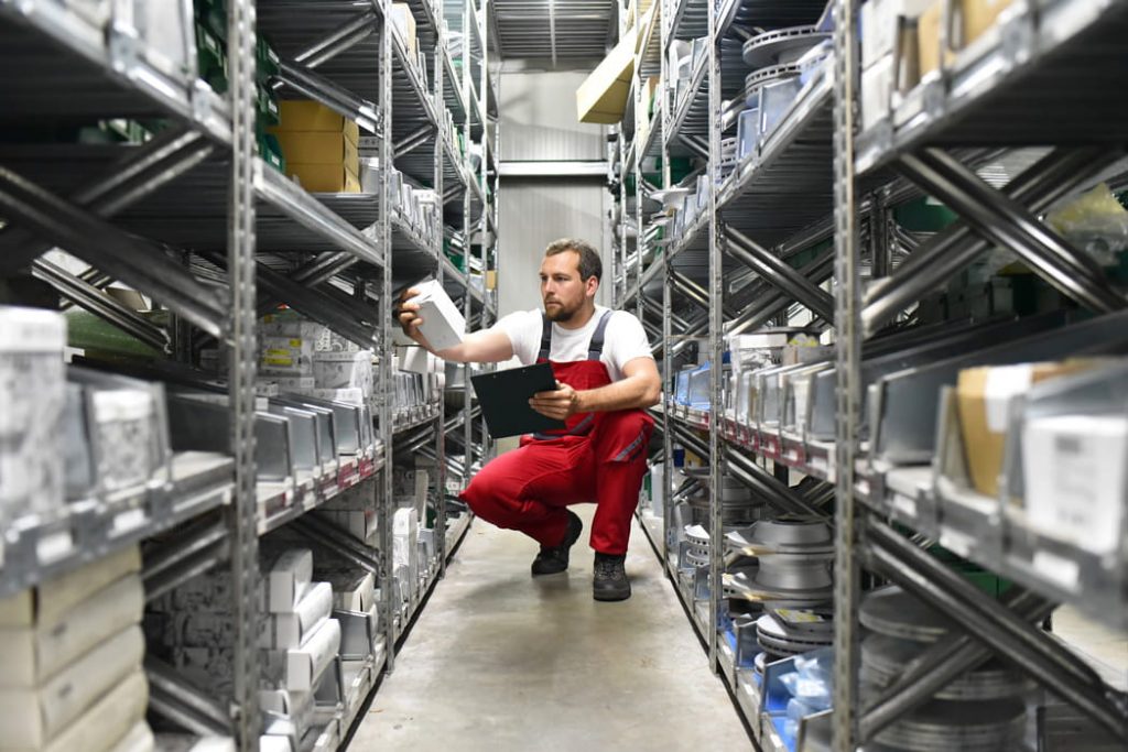 Lagerarbeiter in roter Latzhose entnimmt Artikel aus Metallregal und hält Klemmbrett.