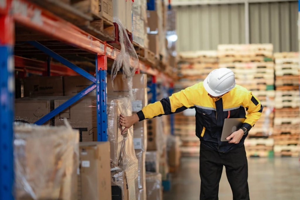 Lagerarbeiter in gelber Schutzjacke und Helm greift in Palettenregal und hält Laptop.