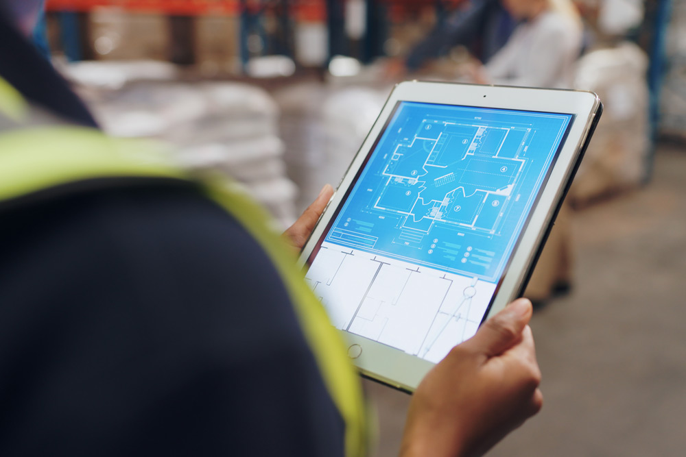 Lagermitarbeiterin in Warnweste hält Tablet mit digitalem Gebäudegrundriss in Lagerhalle.