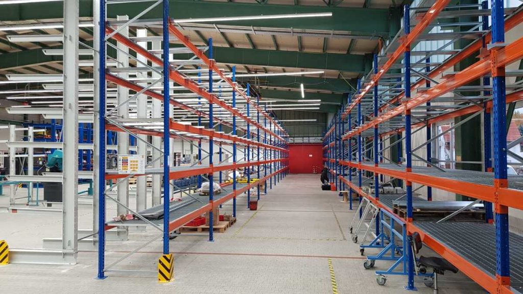 Reihen von Palettenregalen in einer Lagerhalle mit blauen Stützen und orangefarbenen Traversen, teilweise mit Gitterrostböden, im Gang stehen Rollwagen und Transportequipment, Warnmarkierungen am Boden.